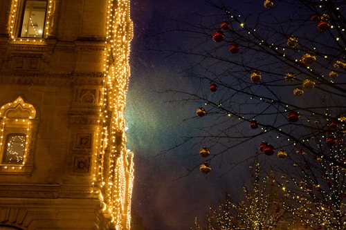 A beautifully decorated building facade is illuminated with golden lights. To the right, a tree is adorned with red and gold ornaments, with its branches outlined by small white lights. The background features a deep blue night sky, enhancing the festive atmosphere.