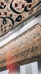 A piece of textured fabric featuring intricately woven and scripted Arabic calligraphy with an artistic and traditional design. There are elements hanging vertically from the fabric, appearing like fine threads or strings.