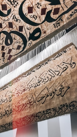 A piece of textured fabric featuring intricately woven and scripted Arabic calligraphy with an artistic and traditional design. There are elements hanging vertically from the fabric, appearing like fine threads or strings.