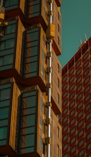 A modern architectural design featuring a glass-covered building with a series of cuboid projections. The structure showcases a copper or bronze frame contrasting with the clear glass panes. To the right, there is another building with a red grid pattern.