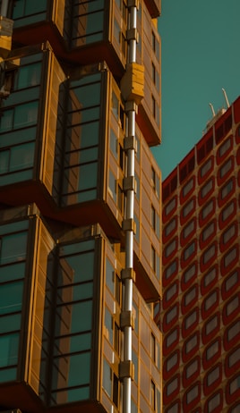 A modern architectural design featuring a glass-covered building with a series of cuboid projections. The structure showcases a copper or bronze frame contrasting with the clear glass panes. To the right, there is another building with a red grid pattern.