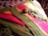 A colorful crochet blanket with stripes of green, pink, and yellow yarn is spread out on a patterned fabric surface. A ball of green yarn and several crochet hook strands are placed on top of the blanket, suggesting an ongoing crochet project.
