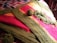 A colorful crochet blanket with stripes of green, pink, and yellow yarn is spread out on a patterned fabric surface. A ball of green yarn and several crochet hook strands are placed on top of the blanket, suggesting an ongoing crochet project.