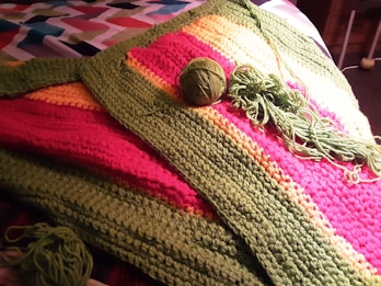A colorful crochet blanket with stripes of green, pink, and yellow yarn is spread out on a patterned fabric surface. A ball of green yarn and several crochet hook strands are placed on top of the blanket, suggesting an ongoing crochet project.