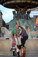 A family poses happily in front of a ornate fountain adorned with classical sculptures. There are two adults and two children, all smiling and wearing winter clothing, including scarves and sunglasses. The setting appears to be in a historic or cultural area, with architectural elements visible in the background.