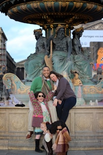 A family poses happily in front of a ornate fountain adorned with classical sculptures. There are two adults and two children, all smiling and wearing winter clothing, including scarves and sunglasses. The setting appears to be in a historic or cultural area, with architectural elements visible in the background.