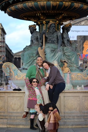A family poses happily in front of a ornate fountain adorned with classical sculptures. There are two adults and two children, all smiling and wearing winter clothing, including scarves and sunglasses. The setting appears to be in a historic or cultural area, with architectural elements visible in the background.