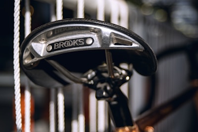 A close-up view of a bicycle seat with the brand name 'Brooks' visible on the metal support under the seat. The background is blurred, showing vertical white ropes or bars, creating a contrast with the metallic and dark colors of the seat.