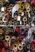A variety of fabric rolls are stacked closely together, each with unique colors and patterns. The rolls are wrapped tightly on cardboard tubes, with some tags attached to them, possibly indicating pricing or fabric details. The fabrics display a range of textures and designs, from plain colors to intricate patterns.