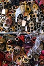 A variety of fabric rolls are stacked closely together, each with unique colors and patterns. The rolls are wrapped tightly on cardboard tubes, with some tags attached to them, possibly indicating pricing or fabric details. The fabrics display a range of textures and designs, from plain colors to intricate patterns.