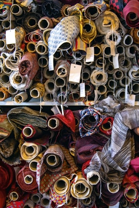 A variety of fabric rolls are stacked closely together, each with unique colors and patterns. The rolls are wrapped tightly on cardboard tubes, with some tags attached to them, possibly indicating pricing or fabric details. The fabrics display a range of textures and designs, from plain colors to intricate patterns.