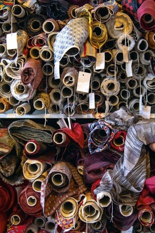 A variety of fabric rolls are stacked closely together, each with unique colors and patterns. The rolls are wrapped tightly on cardboard tubes, with some tags attached to them, possibly indicating pricing or fabric details. The fabrics display a range of textures and designs, from plain colors to intricate patterns.