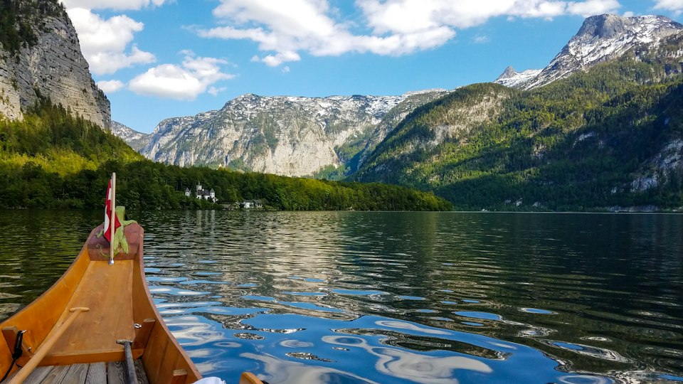 A wooden rowboat crossing a still alpine lake, with forested slopes and snow-dusted peaks in the background