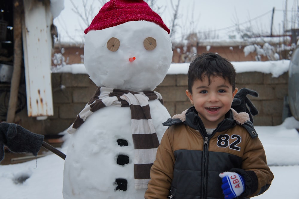 boy in black jacket standing beside snowman during daytime