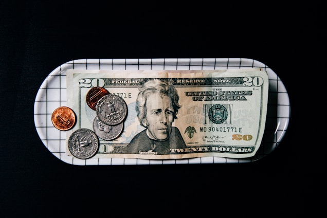 A twenty-dollar bill is placed on a rectangular tray with a grid pattern. Several coins, including quarters, a nickel, and pennies, are scattered across the bill and tray. The background is entirely black, emphasizing the currency and tray.