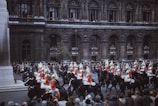 A procession of horse-mounted guards in red uniforms with helmets adorned with white plumes rides past a historic, ornate building. The crowd of spectators watches from the sidewalk and from the building's balconies.