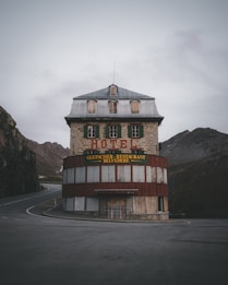 A rustic hotel building with a stone facade and a metal roof, situated at a curve in a mountain road. The hotel features wooden shutters on the windows and a prominent sign that reads 'Hotel' and 'Gletscher - Restaurant Belvedere'. The surroundings include rugged mountain terrain and overcast skies.
