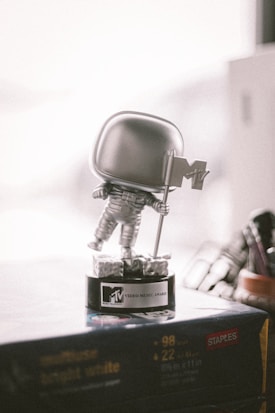 A silver astronaut figurine holding a flag stands on a black base labeled MTV Video Music Award. The scene is lit softly, with a blurred background that includes part of a workspace or shelf.