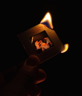 A hand holds a photographic slide that is partially engulfed in flames against a dark background. The image on the slide depicts three individuals engaged in a seemingly casual interaction.