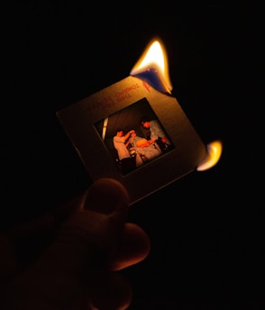 A hand holds a photographic slide that is partially engulfed in flames against a dark background. The image on the slide depicts three individuals engaged in a seemingly casual interaction.