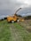 A large yellow agricultural machine, specifically a New Holland forage harvester, is parked on a grassy field next to a cornfield. The machine is equipped with a long arm for cutting and processing crops. The sky is overcast, suggesting an impending storm or cloudy weather. The earth is damp, with visible tire tracks leading to the harvester, indicating recent use.