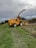A large yellow agricultural machine, specifically a New Holland forage harvester, is parked on a grassy field next to a cornfield. The machine is equipped with a long arm for cutting and processing crops. The sky is overcast, suggesting an impending storm or cloudy weather. The earth is damp, with visible tire tracks leading to the harvester, indicating recent use.
