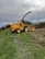 A large yellow agricultural machine, specifically a New Holland forage harvester, is parked on a grassy field next to a cornfield. The machine is equipped with a long arm for cutting and processing crops. The sky is overcast, suggesting an impending storm or cloudy weather. The earth is damp, with visible tire tracks leading to the harvester, indicating recent use.