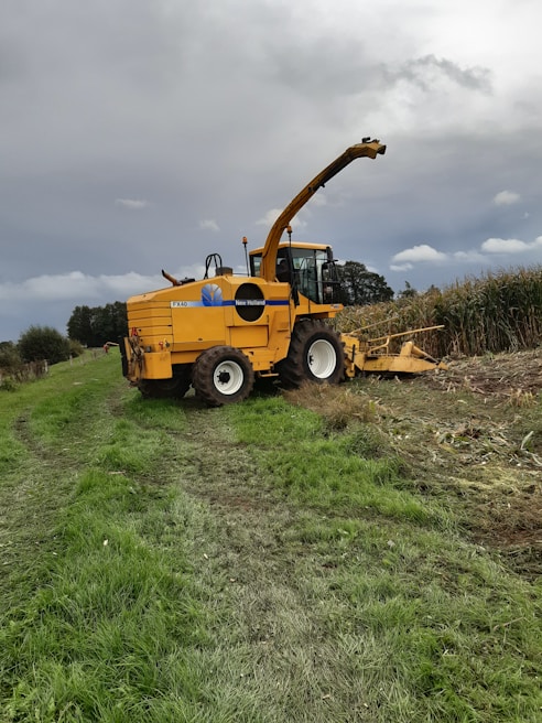 A large yellow agricultural machine, specifically a New Holland forage harvester, is parked on a grassy field next to a cornfield. The machine is equipped with a long arm for cutting and processing crops. The sky is overcast, suggesting an impending storm or cloudy weather. The earth is damp, with visible tire tracks leading to the harvester, indicating recent use.