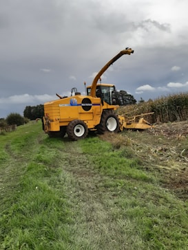 A large yellow agricultural machine, specifically a New Holland forage harvester, is parked on a grassy field next to a cornfield. The machine is equipped with a long arm for cutting and processing crops. The sky is overcast, suggesting an impending storm or cloudy weather. The earth is damp, with visible tire tracks leading to the harvester, indicating recent use.