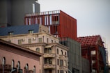 A modern urban scene featuring a combination of architectural styles. In the foreground, there is a building with classical design elements, such as decorative molding and arched windows. Adjacent to it are more contemporary structures with red and gray facades, including a building with a red lattice framework.
