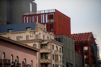 A modern urban scene featuring a combination of architectural styles. In the foreground, there is a building with classical design elements, such as decorative molding and arched windows. Adjacent to it are more contemporary structures with red and gray facades, including a building with a red lattice framework.