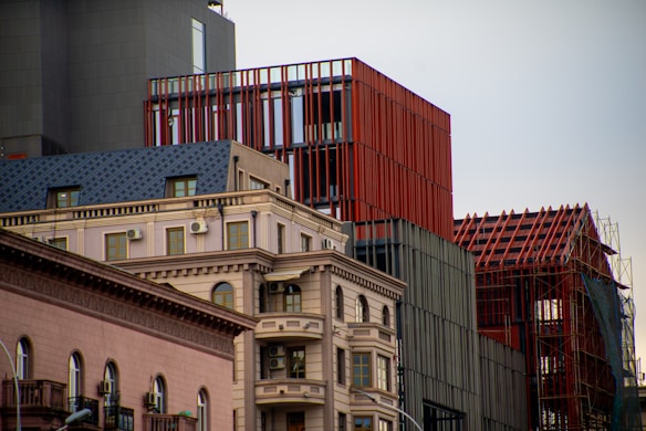 A modern urban scene featuring a combination of architectural styles. In the foreground, there is a building with classical design elements, such as decorative molding and arched windows. Adjacent to it are more contemporary structures with red and gray facades, including a building with a red lattice framework.