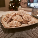 Several cookies dusted with powdered sugar are arranged in a pile on a decorative plate. In the background, there are blurry holiday lights and a clock visible, suggesting a cozy and festive setting.