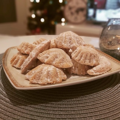 Several cookies dusted with powdered sugar are arranged in a pile on a decorative plate. In the background, there are blurry holiday lights and a clock visible, suggesting a cozy and festive setting.