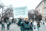 A group of people is participating in a protest, holding signs related to an abortion issue. The woman in the forefront is smiling and wearing a yellow beanie and a dark jacket. The crowd is gathered near a street, with a building and bare trees visible in the background, suggesting it's a cool day.