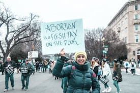 A group of people is participating in a protest, holding signs related to an abortion issue. The woman in the forefront is smiling and wearing a yellow beanie and a dark jacket. The crowd is gathered near a street, with a building and bare trees visible in the background, suggesting it's a cool day.