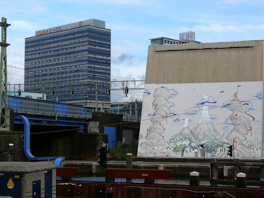 A large, modern building with the sign 'Mövenpick Hotel' is positioned next to a bridge with blue structural elements. In the foreground, there is a wall with an artistic mural depicting abstract mountain and cloud formations, possibly inspired by Asian art. A railway structure with overhead wires is visible, contributing to an urban setting.