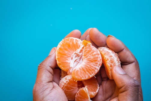Hands delicately holding peeled slices of a juicy orange against a bright blue background.