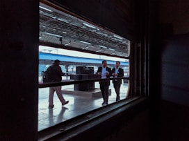 A view from inside a train looking out onto a platform where several people are visible. Two men in business attire are walking together, engaged in conversation. Another person is walking alone in the opposite direction, carrying a backpack. The platform is partially covered with a blue roof, and metal beams and lighting fixtures are visible.