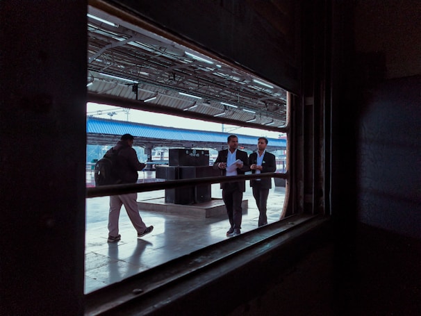 A view from inside a train looking out onto a platform where several people are visible. Two men in business attire are walking together, engaged in conversation. Another person is walking alone in the opposite direction, carrying a backpack. The platform is partially covered with a blue roof, and metal beams and lighting fixtures are visible.