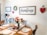 A cozy dining room with a rustic farmhouse theme, showcasing a wooden table set for a meal with plates and napkins. A bouquet of red and white roses in a vase and a dome-covered plate of muffins sit on the table. The walls are adorned with farmhouse-style decorations including a wreath, signs, and a heart-shaped wall hanging. A small cart holds a coffee maker and mugs.