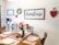 A cozy dining room with a rustic farmhouse theme, showcasing a wooden table set for a meal with plates and napkins. A bouquet of red and white roses in a vase and a dome-covered plate of muffins sit on the table. The walls are adorned with farmhouse-style decorations including a wreath, signs, and a heart-shaped wall hanging. A small cart holds a coffee maker and mugs.