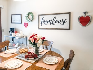 A cozy dining room with a rustic farmhouse theme, showcasing a wooden table set for a meal with plates and napkins. A bouquet of red and white roses in a vase and a dome-covered plate of muffins sit on the table. The walls are adorned with farmhouse-style decorations including a wreath, signs, and a heart-shaped wall hanging. A small cart holds a coffee maker and mugs.