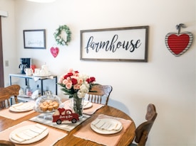 A cozy dining room with a rustic farmhouse theme, showcasing a wooden table set for a meal with plates and napkins. A bouquet of red and white roses in a vase and a dome-covered plate of muffins sit on the table. The walls are adorned with farmhouse-style decorations including a wreath, signs, and a heart-shaped wall hanging. A small cart holds a coffee maker and mugs.