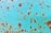 A close-up view of a rusted metal surface with a turquoise-colored paint that is peeling and worn. Brown rust patches are scattered across the surface, creating an aged and textured appearance.