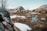 A rustic cabin is nestled in a mountainous landscape, surrounded by snow-covered ground and rocky terrain. In the foreground, a small stream winds through the area, providing a sense of tranquility. Bare trees are scattered around, adding to the serene, wintery setting.