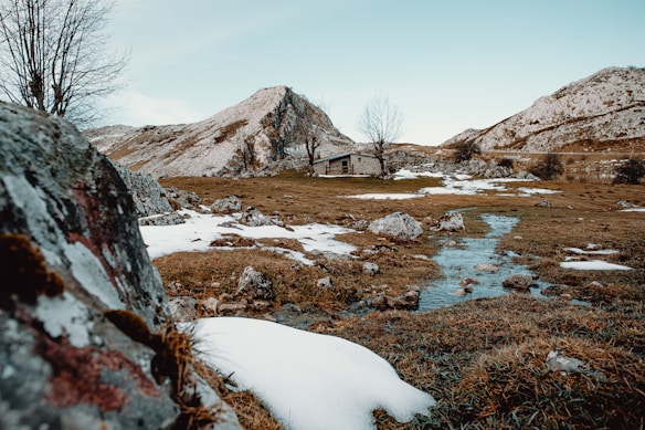 A rustic cabin is nestled in a mountainous landscape, surrounded by snow-covered ground and rocky terrain. In the foreground, a small stream winds through the area, providing a sense of tranquility. Bare trees are scattered around, adding to the serene, wintery setting.