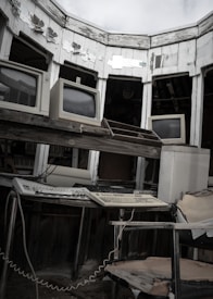 A collection of vintage computer monitors and keyboards is arranged within a decrepit and weathered room with peeling paint. The dark, worn-out chair in the foreground adds to the abandoned and aged atmosphere.