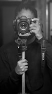 A person holds a camera up to their face, looking directly at the lens. The camera is mounted on a monopod and features a strap with the brand name visible. The image is in black and white, emphasizing the contrast and the details of the camera and the person's hands.