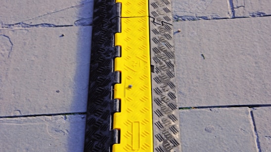 A bright yellow and black cable protector placed on a gray pavement with visible concrete slabs and seams.